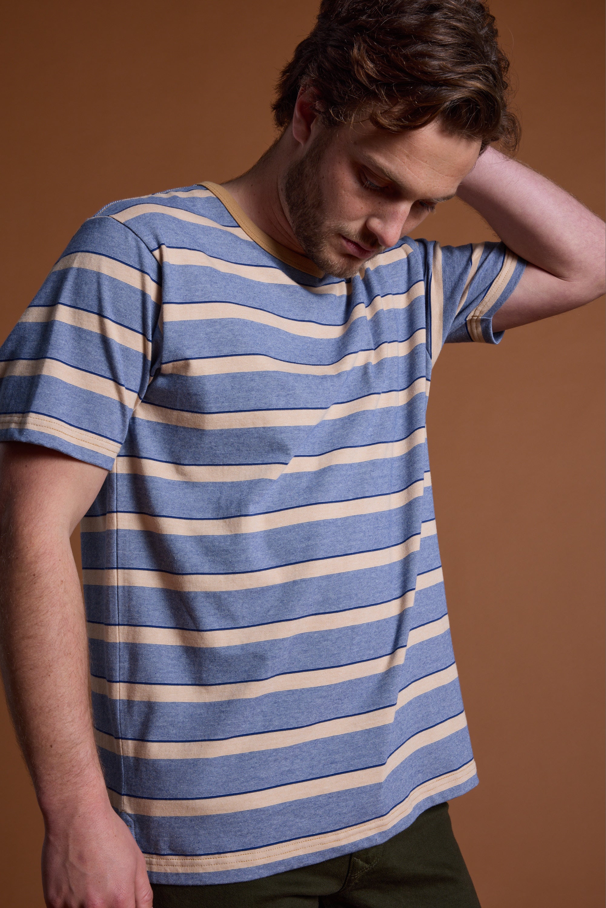 A man wearing the Barkers Chester Stripe Tee in blue and beige stands against a brown background, looking down with one hand on the back of his head.