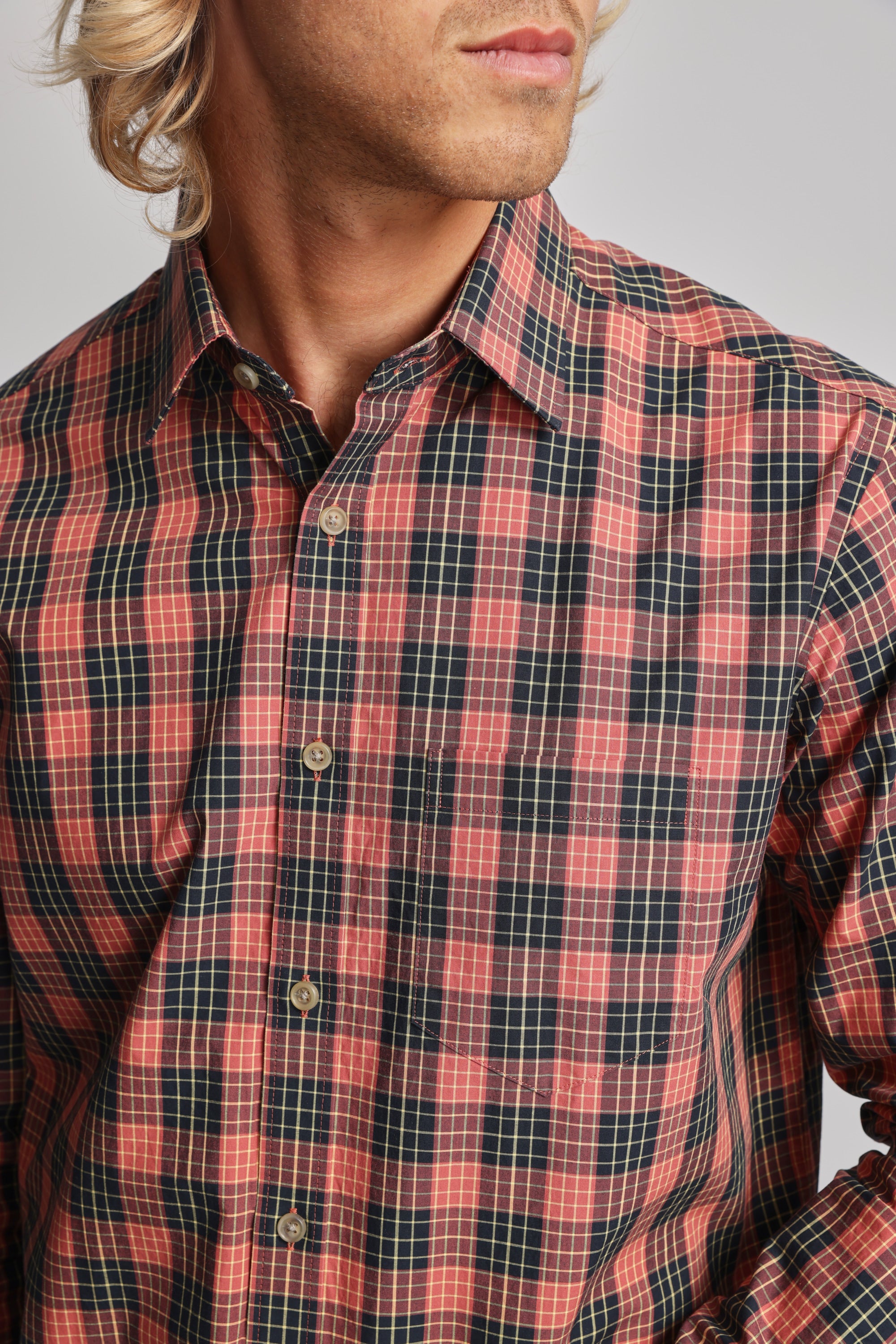 A person with wavy blond hair wears the Barkers Formby Long Sleeve Shirt—a red and black plaid, classic fit style with a classic collar and beige buttons, made from responsibly sourced cotton. Shown from shoulders to above the mouth.