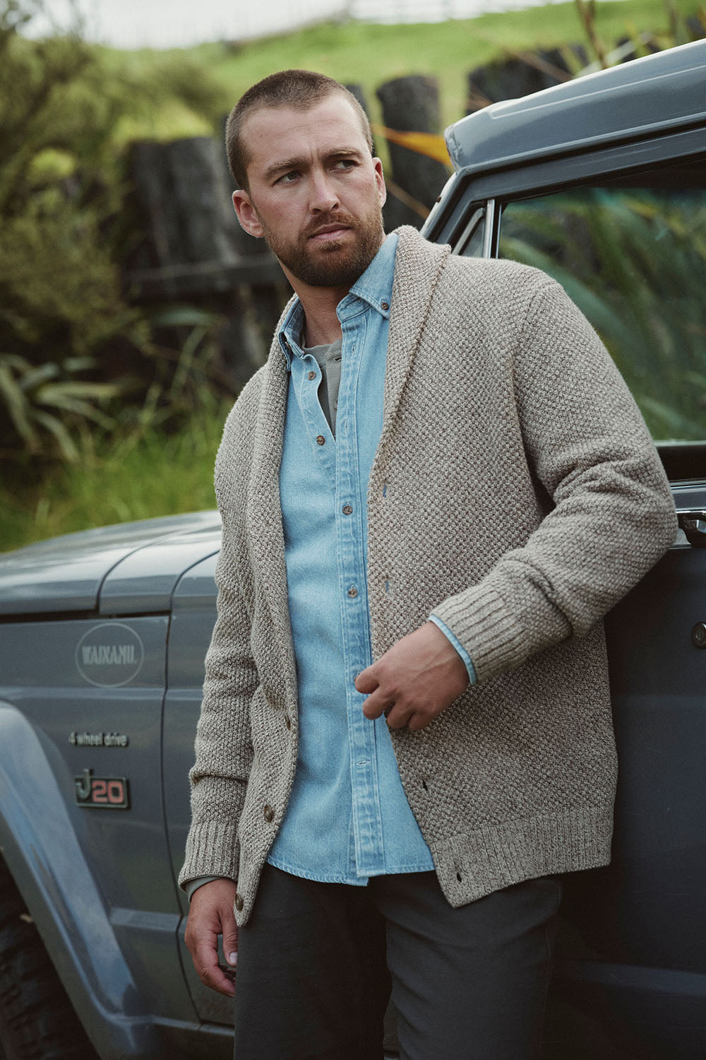 Man in light sweater and denim shirt stands by a gray Jeep in scenic NZ outdoors.