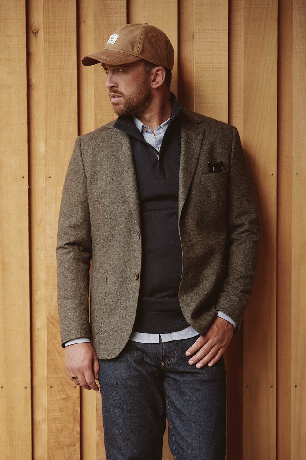Man in a brown blazer, blue sweater, and tan cap in NZ, standing by a wooden wall, looking left.