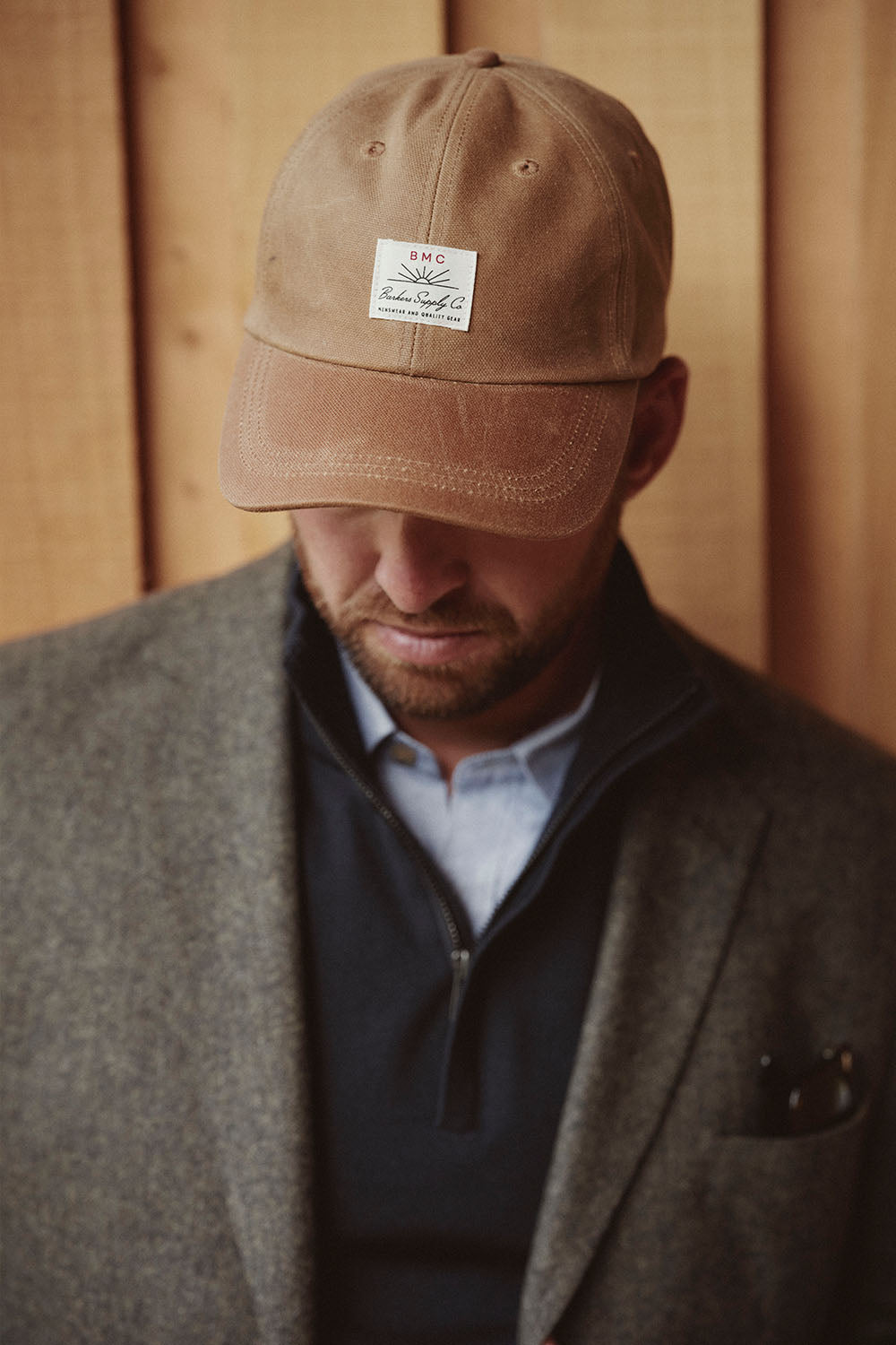 Man in a brown cap and grey blazer looks down, standing against a wooden NZ wooden background.