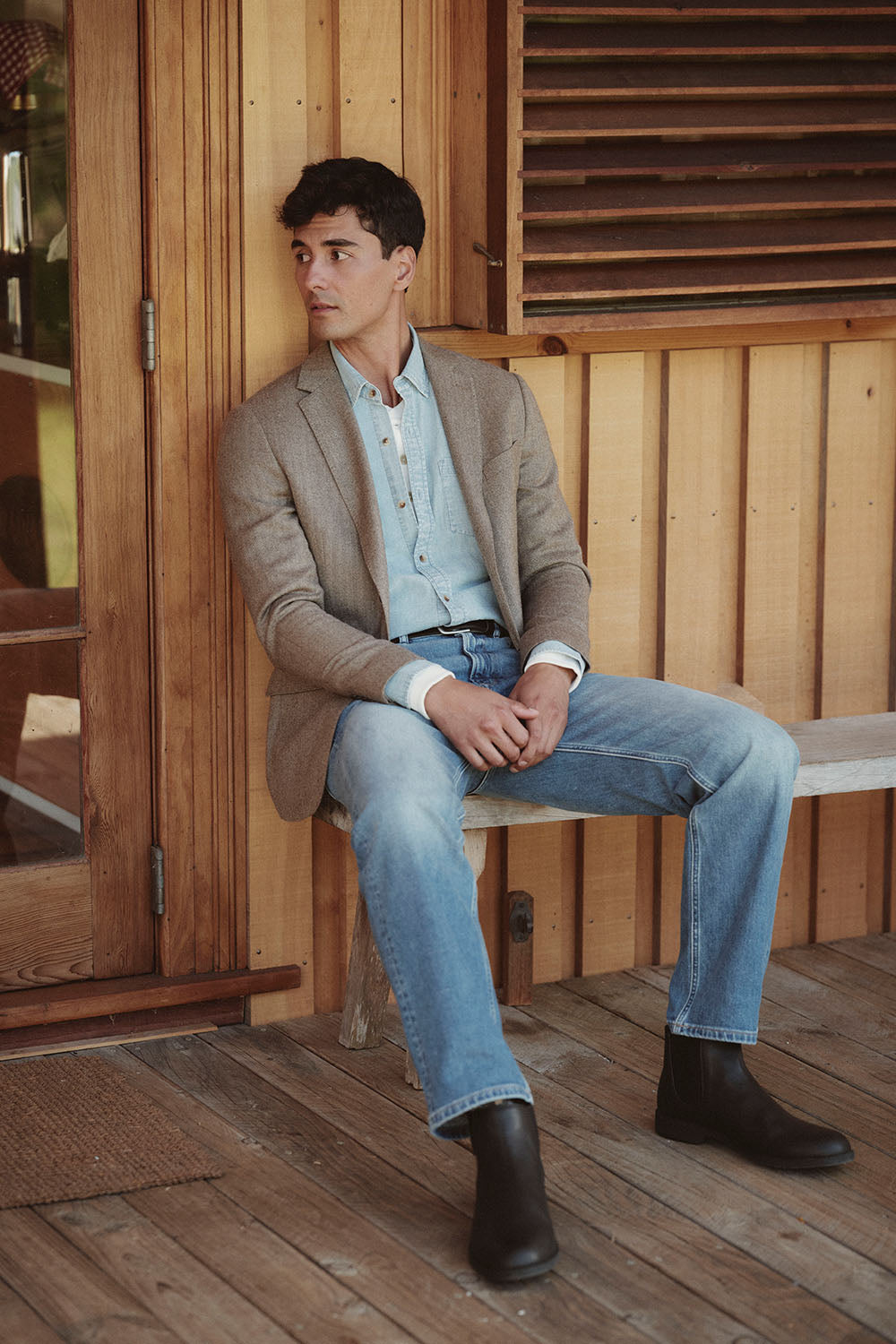 Man in a brown blazer, denim shirt, and jeans sits on a bench by an NZ wooden wall.