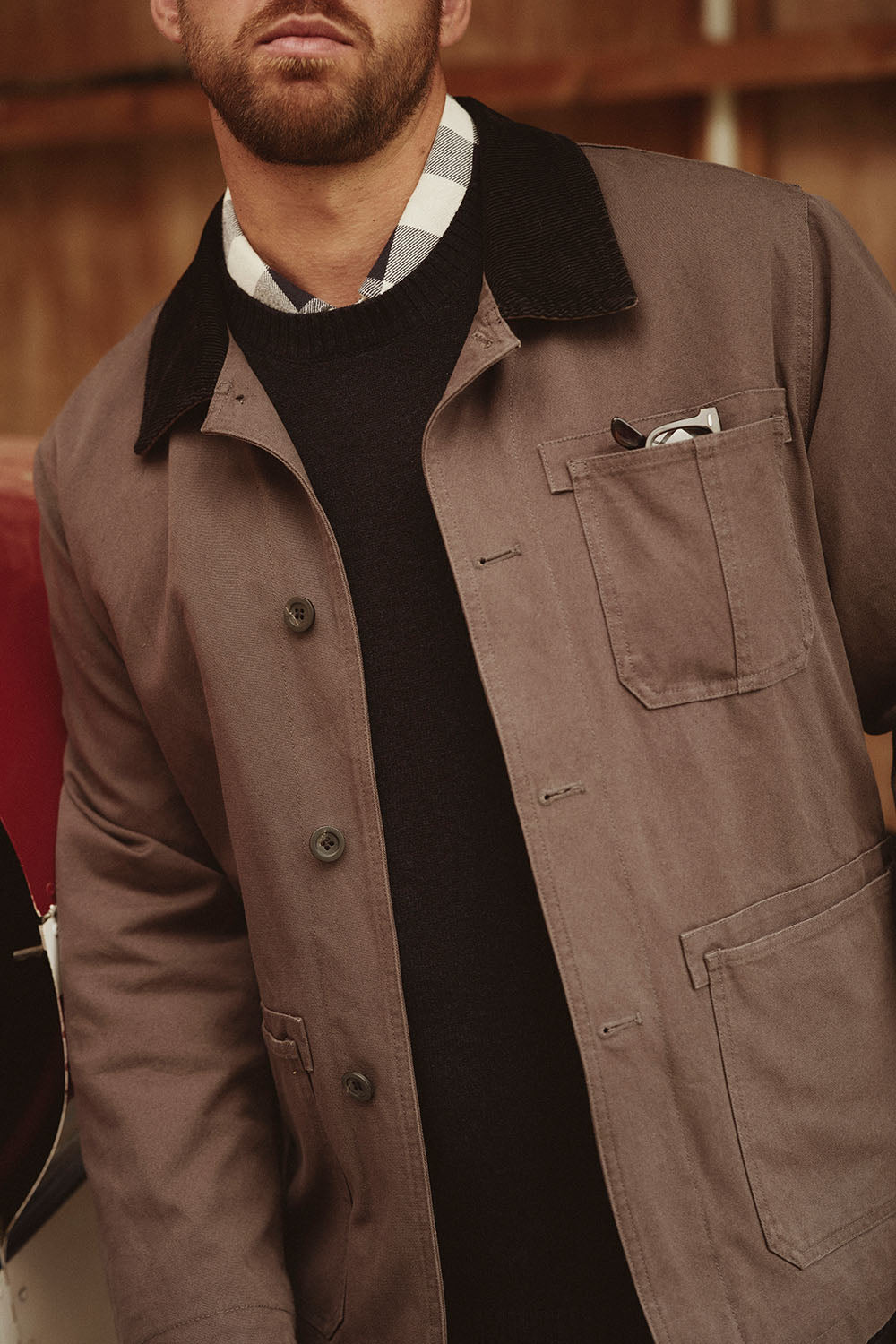 Man wearing a brown jacket with black collar, plaid shirt, NZ style, and black sweater underneath.