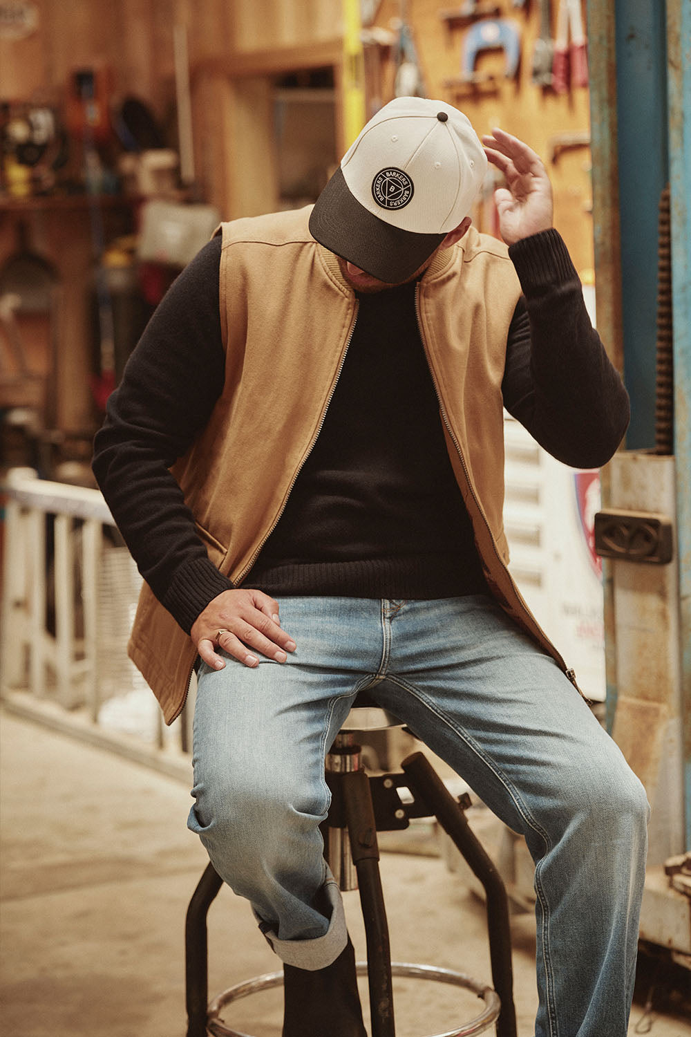Man in a NZ workshop sits on a stool, adjusting his cap, wearing jeans and a tan vest.