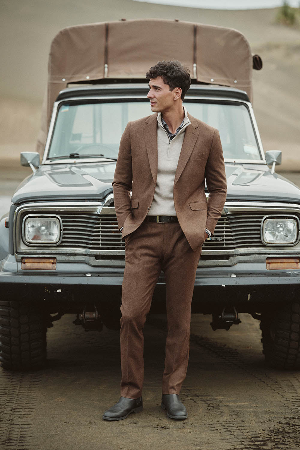 Man in a brown suit stands before a vintage NZ off-road vehicle on a sandy surface.