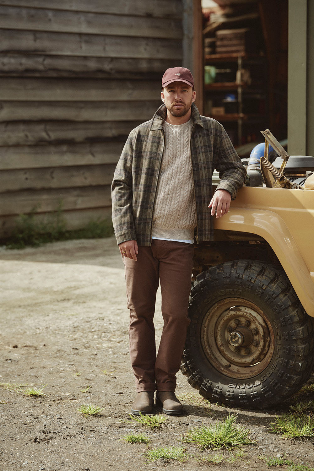 Man in casual fall clothing stands beside a yellow vehicle, with NZ wooden building in background.