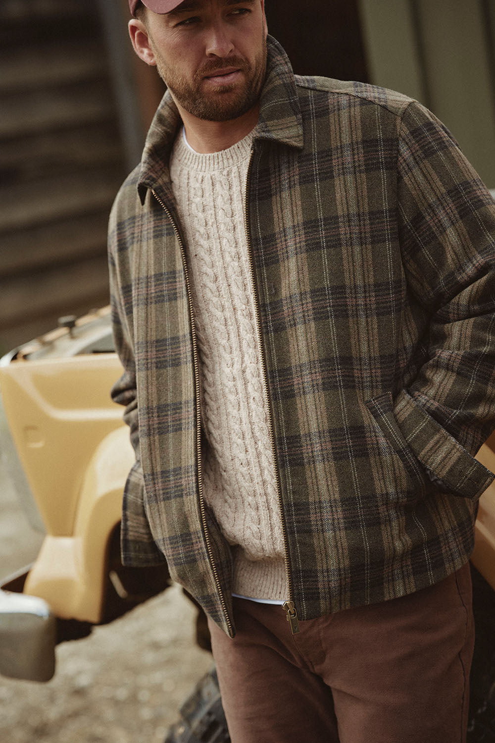 Man in plaid jacket, cream cable-knit sweater, and cap stands by a yellow vehicle in NZ outdoors.