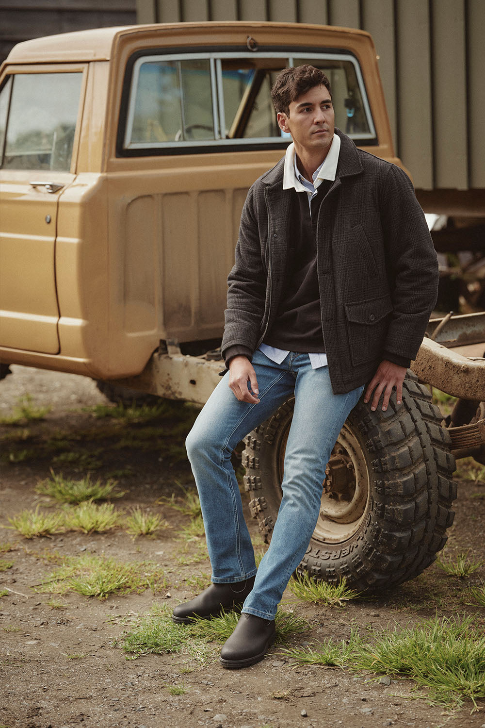 Man in casual clothes sits on a truck tire outdoors, with an old NZ beige truck in the background.