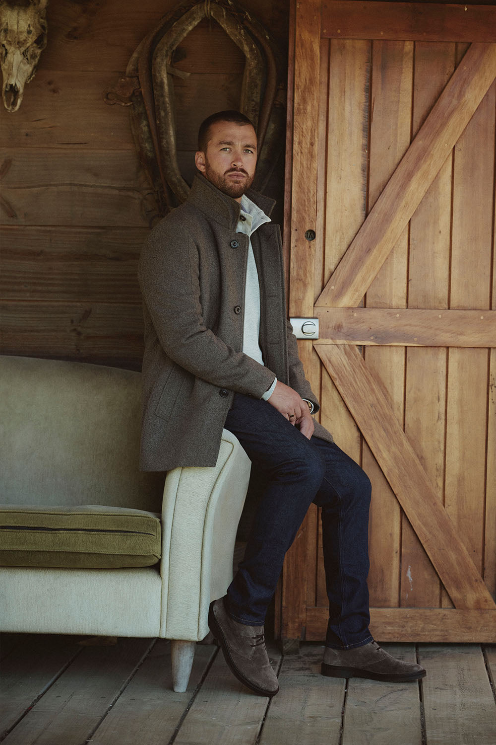 Man in a coat sits on a sofa next to a wooden door in a rustic NZ setting.