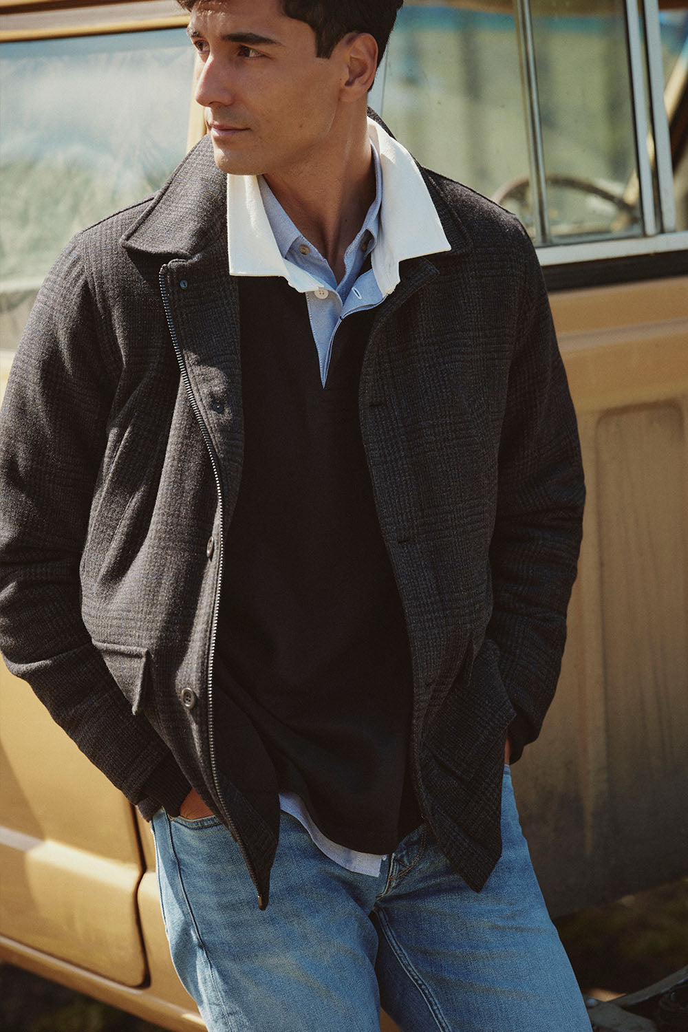Man in layered clothing stands by a tan NZ vehicle, looking to the side with hands in pockets.
