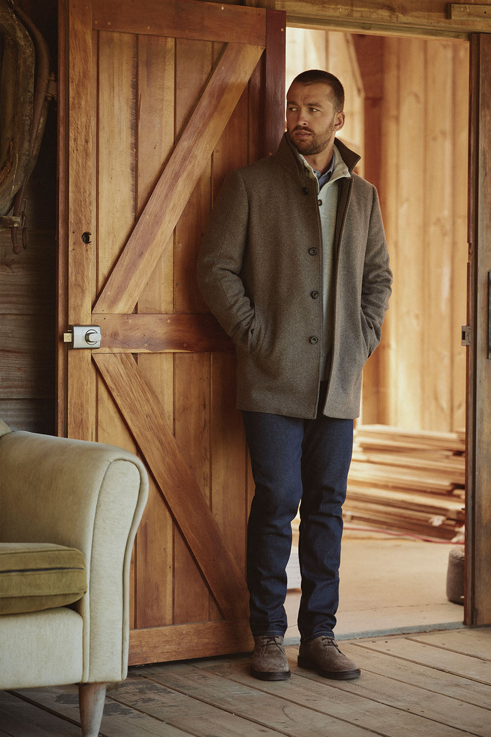 Man in a gray coat stands by a wooden NZ door, hands in pockets, looking to the side.