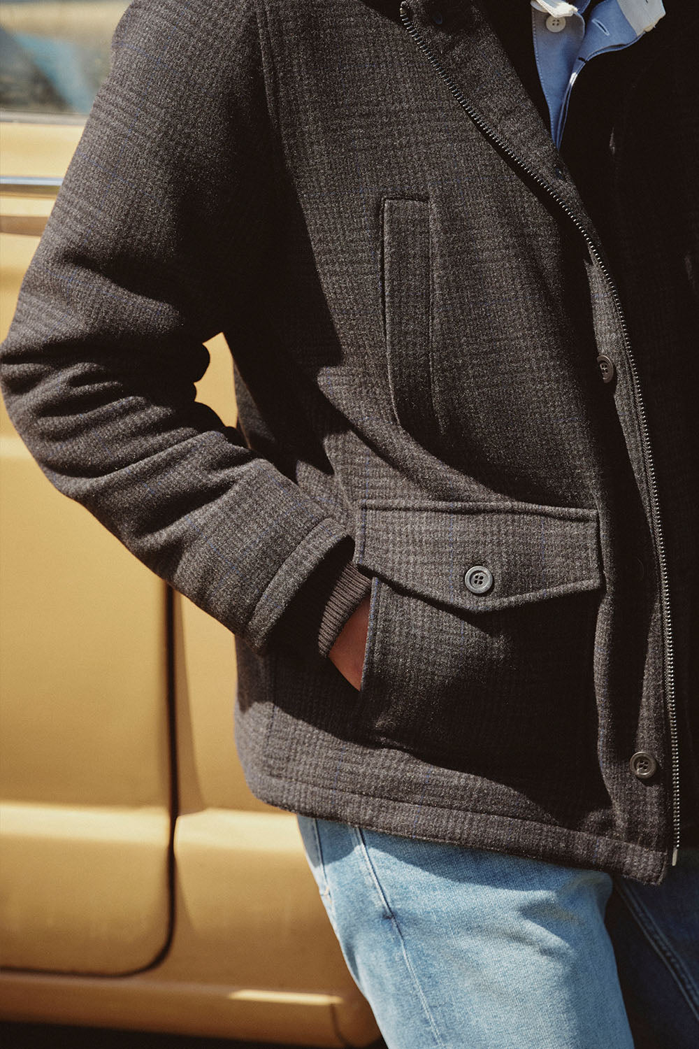 Person in a dark jacket and jeans stands with hand in pocket near a yellow NZ vehicle.