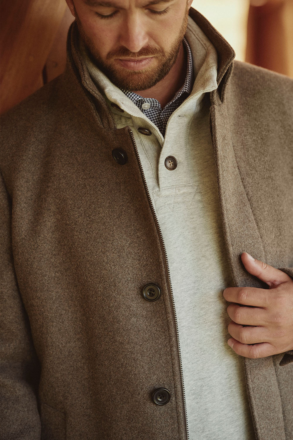 Man wearing a brown coat over a light sweater, looking down in classic NZ style.
