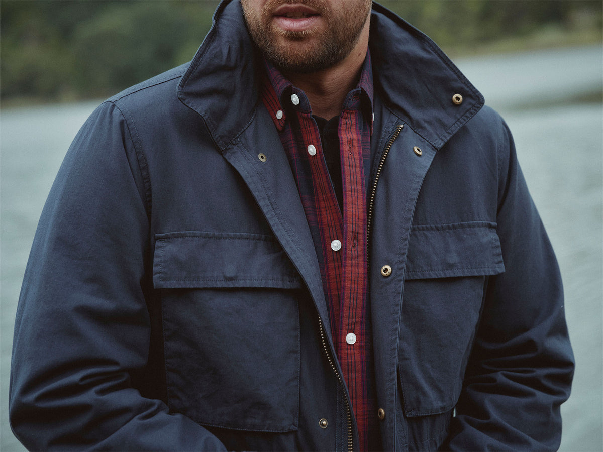 Man in a navy jacket and red plaid shirt standing outdoors by NZ waterfront.