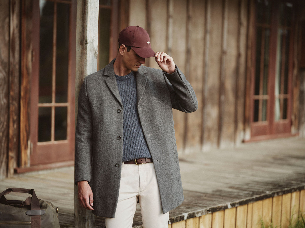 Man in a gray coat and maroon cap stands on a rustic NZ porch, holding his cap brim.