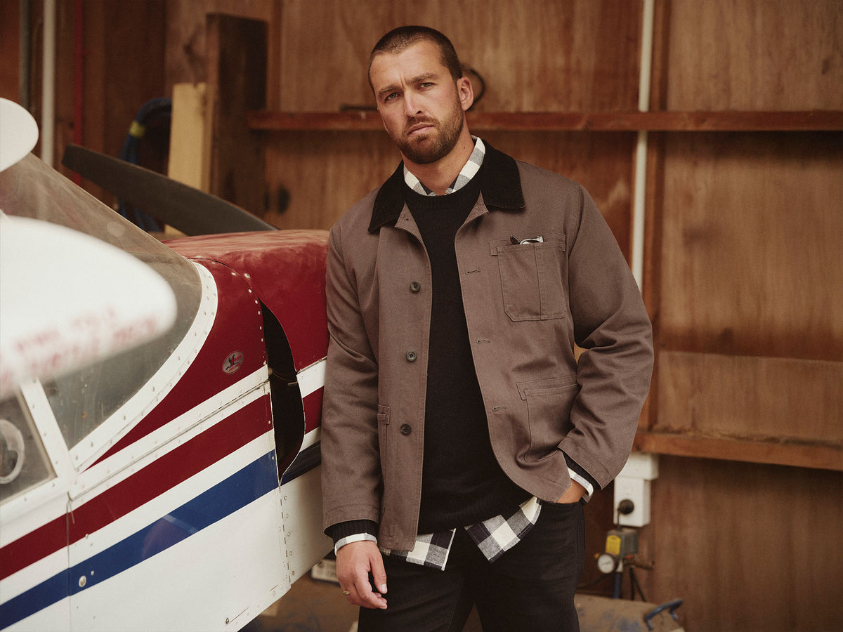 Man in brown jacket stands by an NZ plane inside a wooden hangar, looking at the camera.