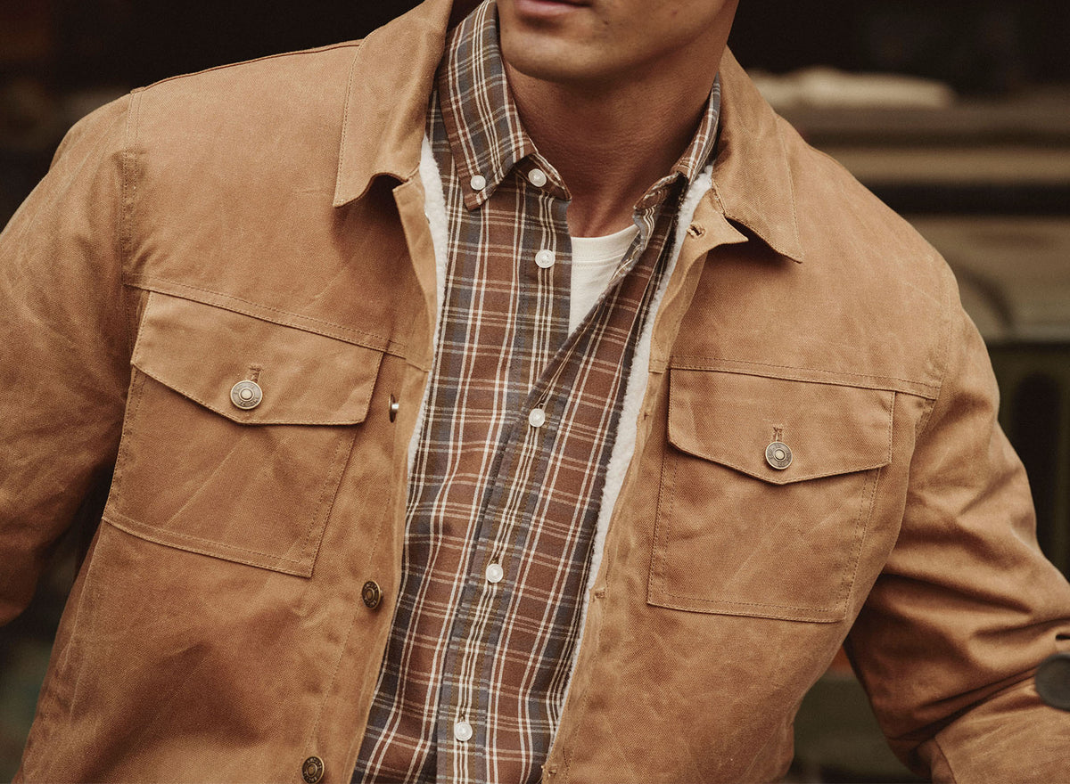 Man wearing a brown jacket over a plaid shirt, NZ style, shown from shoulders to waist.
