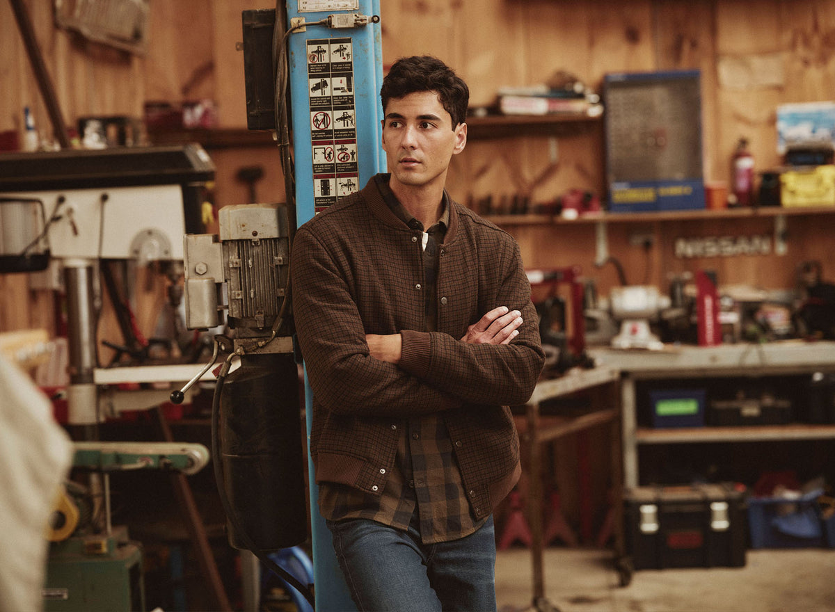 Man in brown jacket stands in a NZ workshop with tools and equipment in the background.
