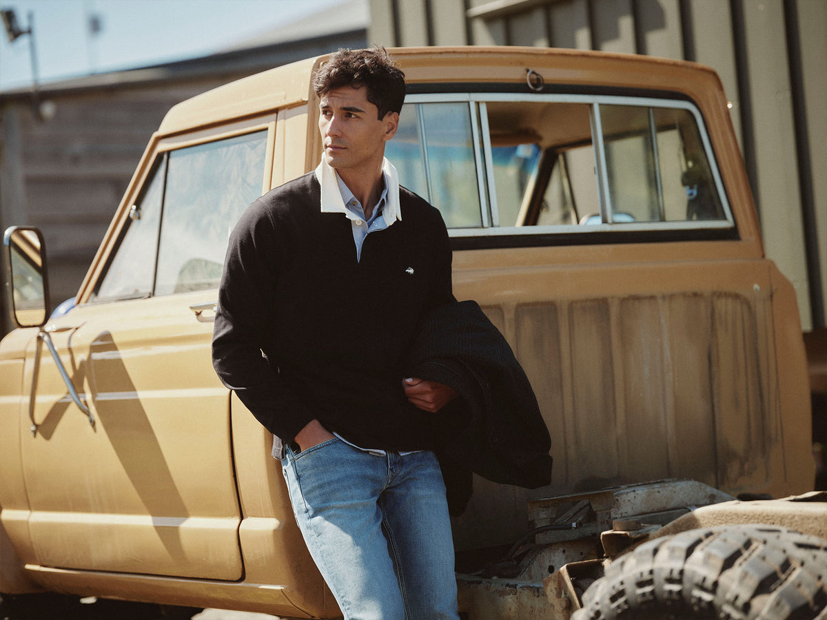 Man in black sweater and jeans leans against a vintage yellow NZ pickup truck outdoors.