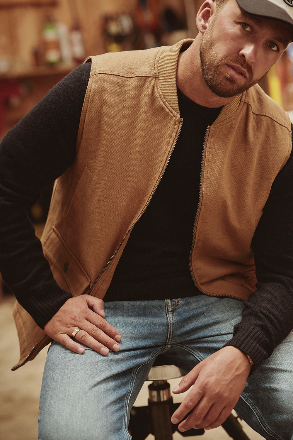 Man in tan vest, black sweater, and jeans sitting indoors, looking at the camera in NZ.