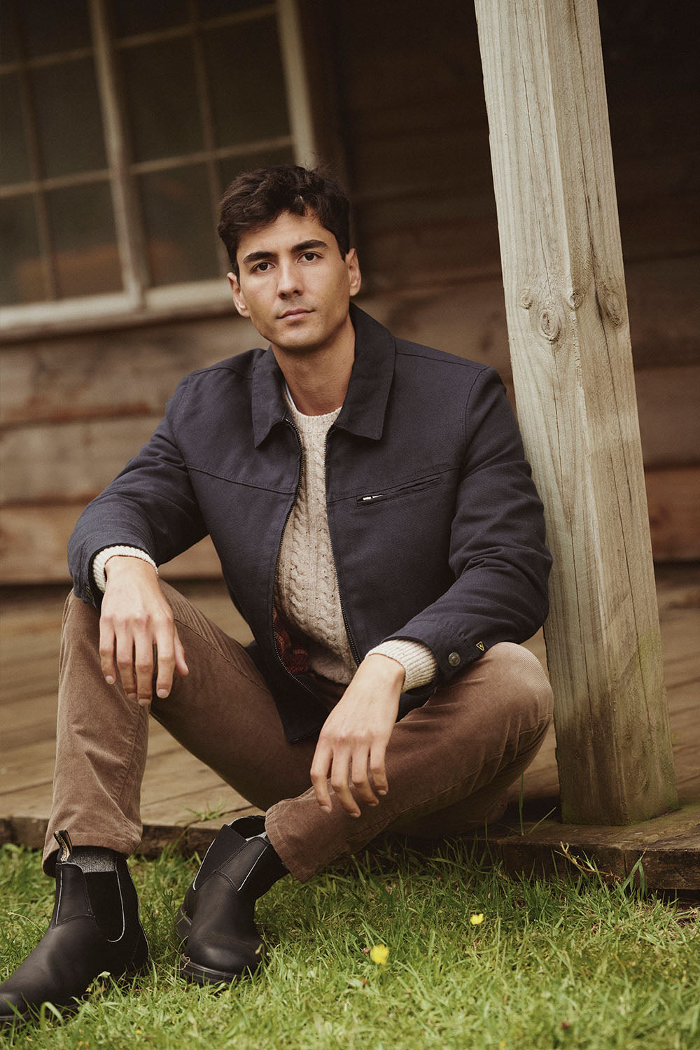 Man sitting on grass by a wooden porch in NZ, wearing a dark jacket, sweater, and brown pants.