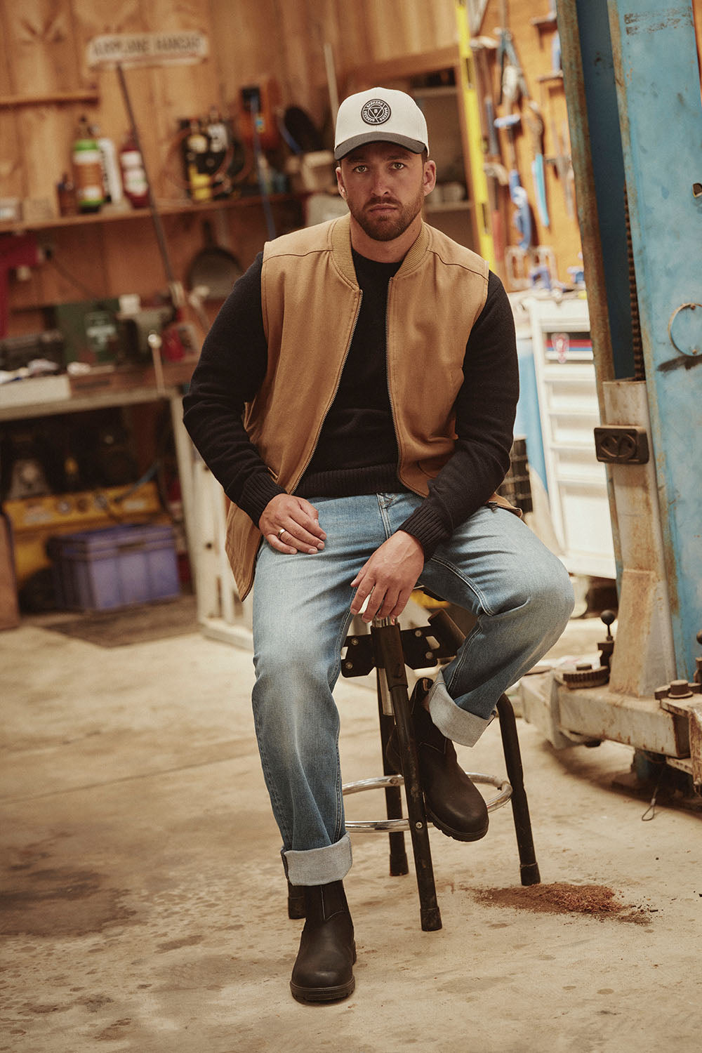 Man in a cap and tan vest sits on a stool in an NZ workshop, looking at the camera.