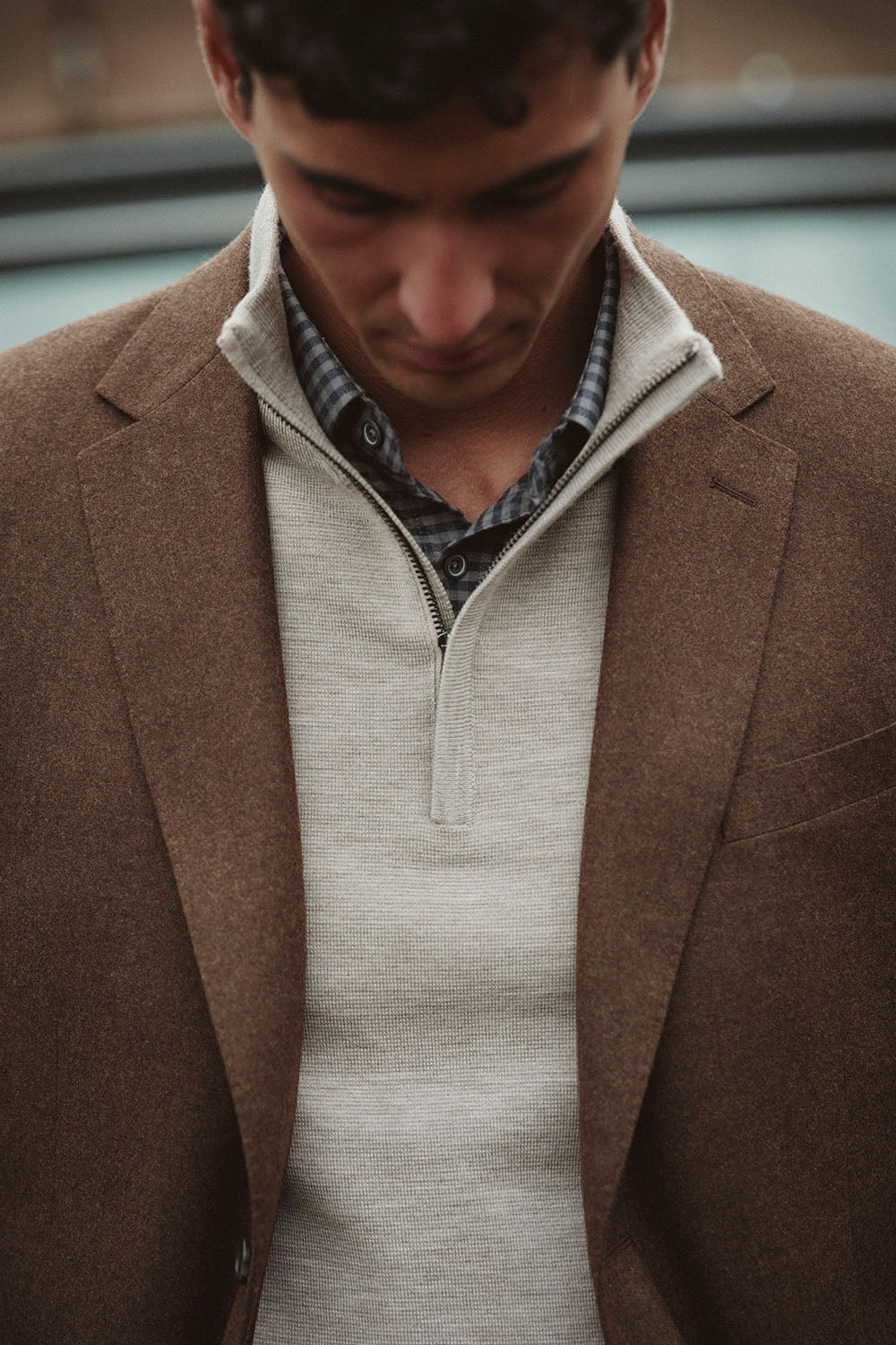 Man looking down, wearing a brown blazer over a light sweater and checked NZ shirt.