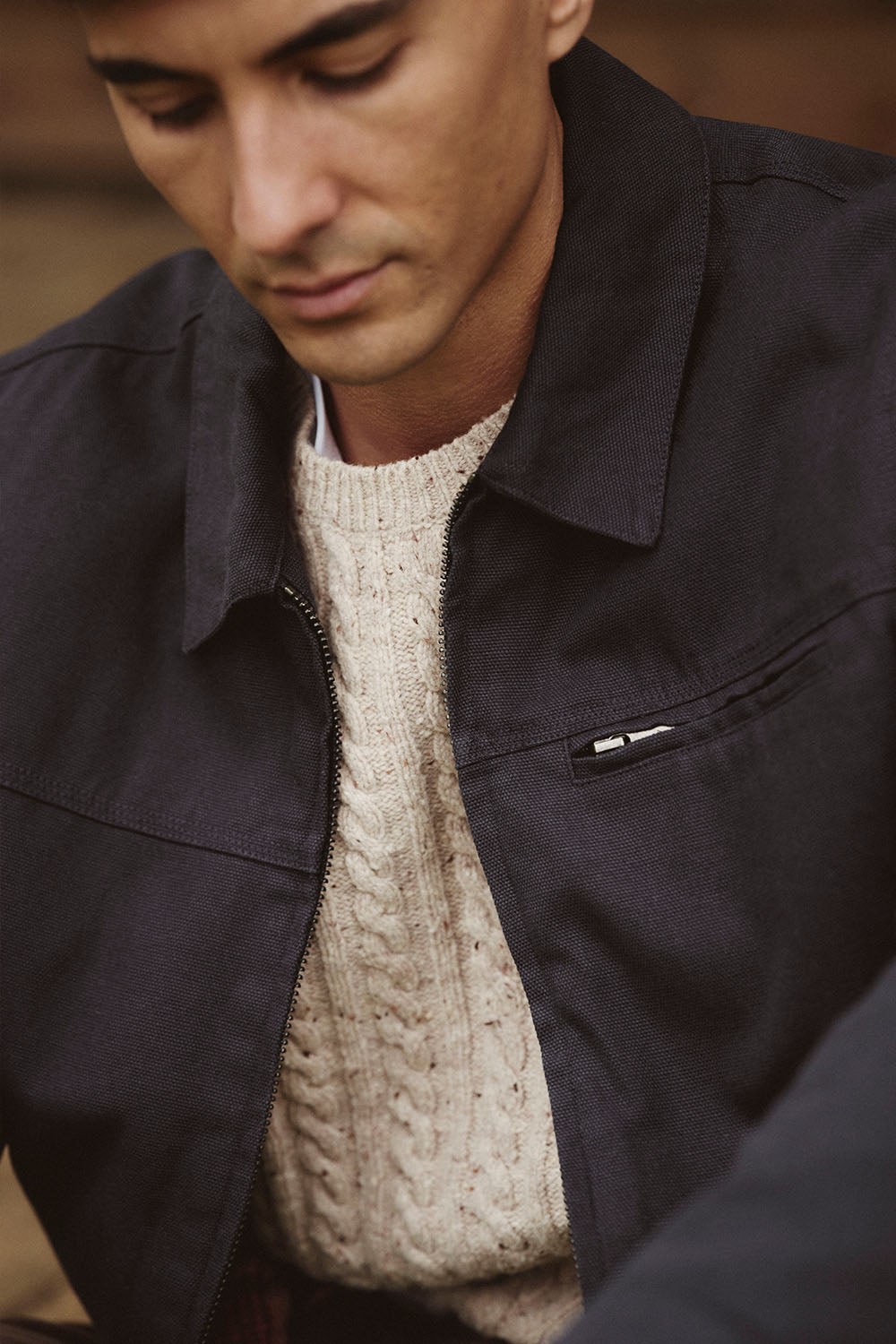 Man wearing a black jacket over a cream cable-knit sweater, looking downward in NZ.