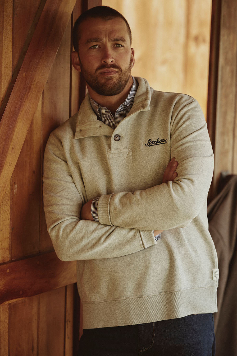 Man in a light gray Packers sweatshirt stands with arms crossed by a wooden NZ wall.