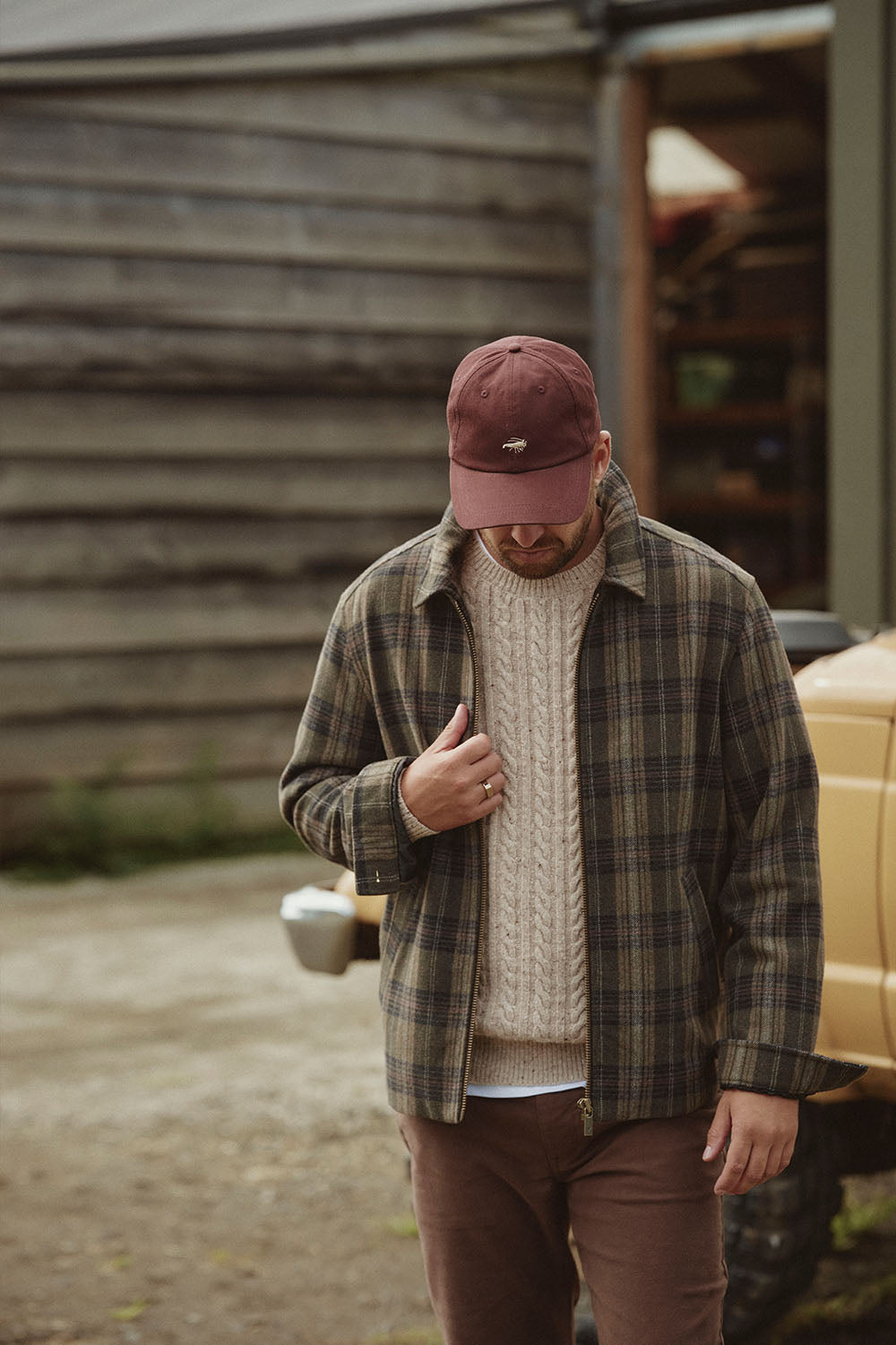 Man in plaid jacket, knit sweater, and cap stands outside by a yellow vehicle in NZ.