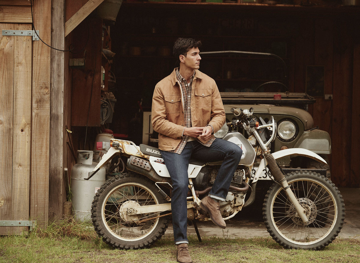 Man in a brown jacket leans on a white dirt bike in front of an NZ wooden garage.