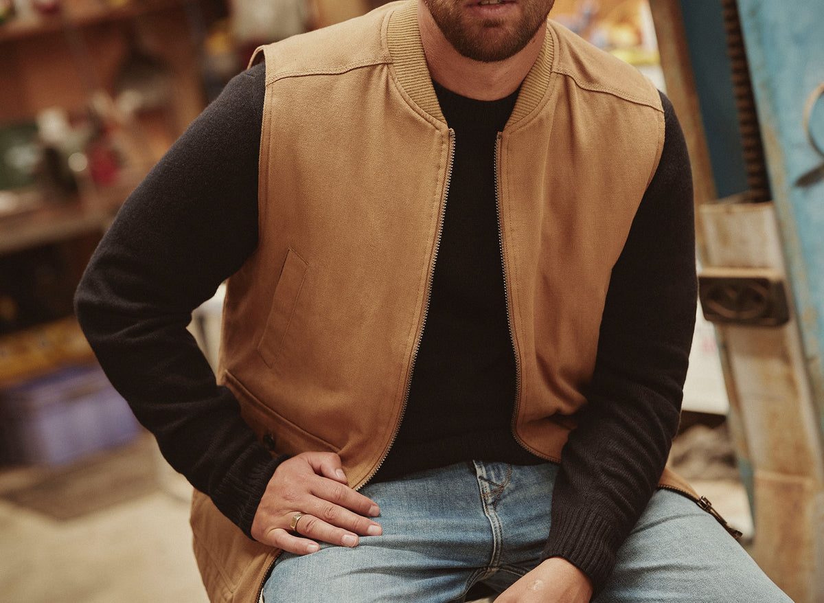 Man wearing a tan vest, black sweater, and jeans sits indoors with an NZ vibe.
