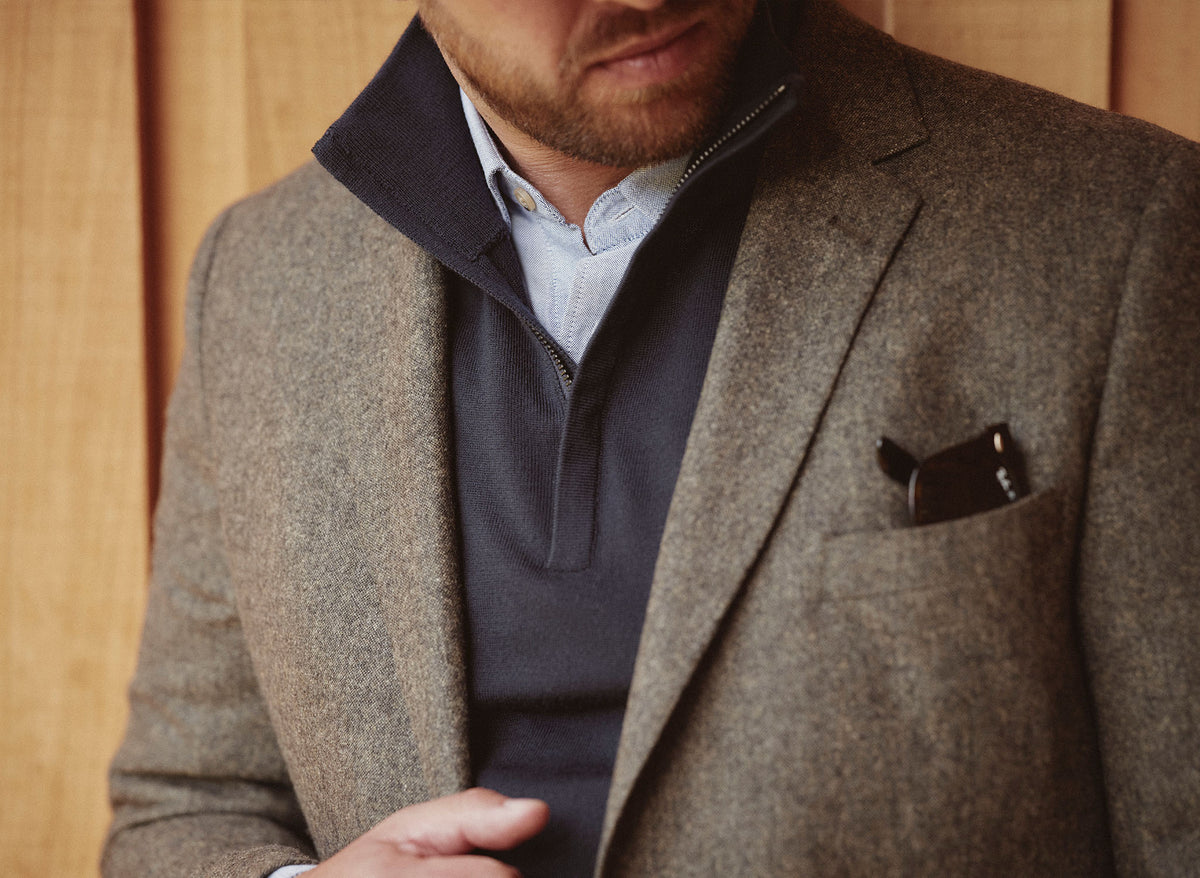 Man in NZ style: brown blazer, navy sweater, blue shirt, sunglasses in pocket.