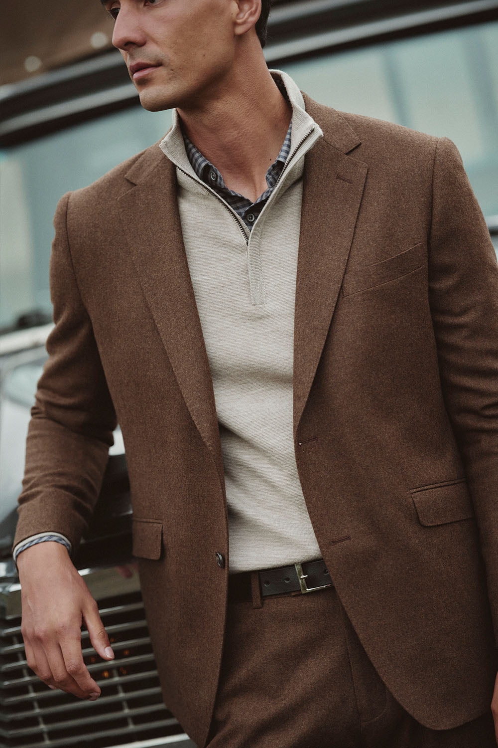 Man wearing a brown suit and beige sweater, standing by a black NZ vehicle.
