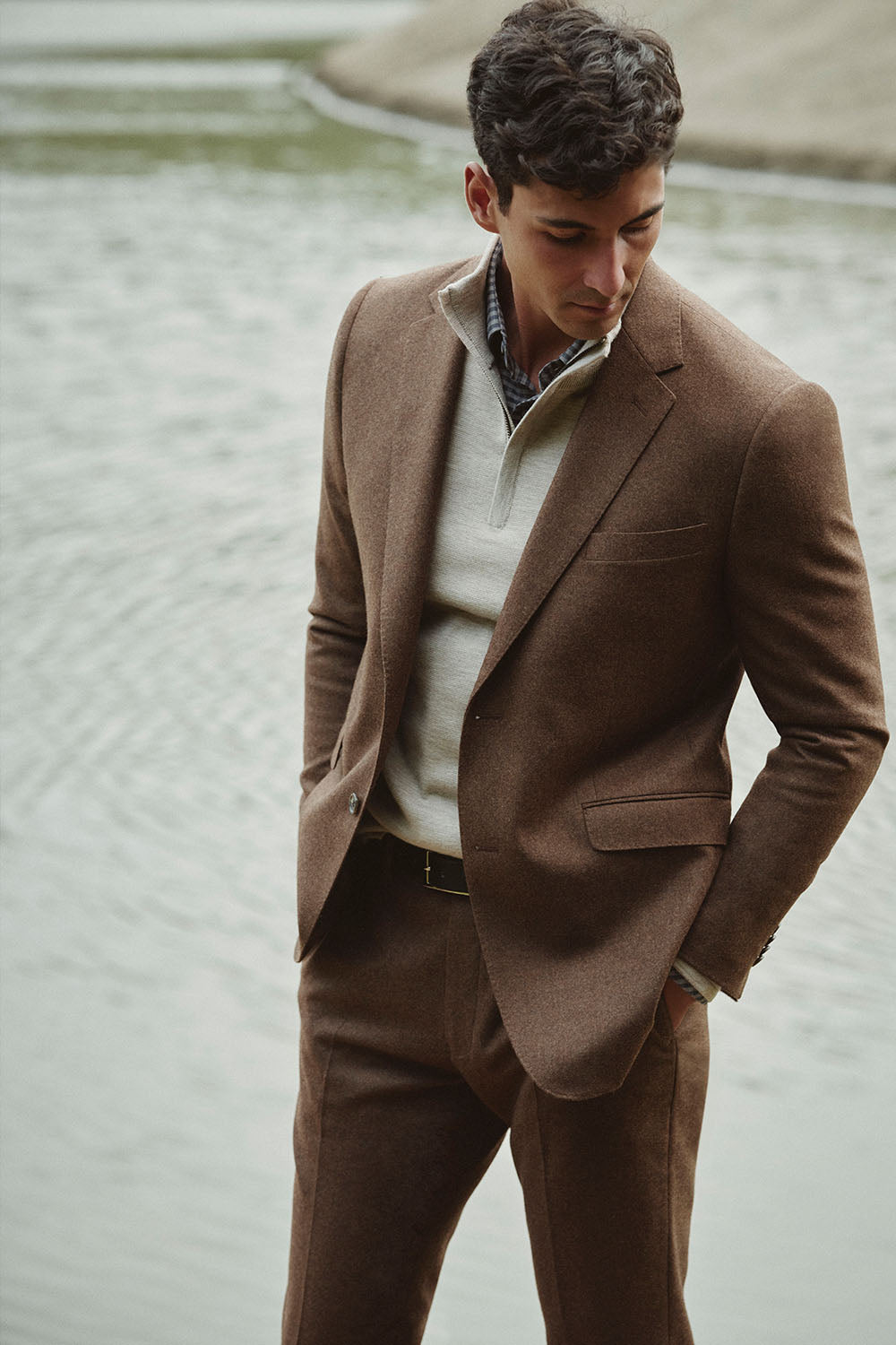 Man in a brown suit by NZ water, looking down with hands in pockets.