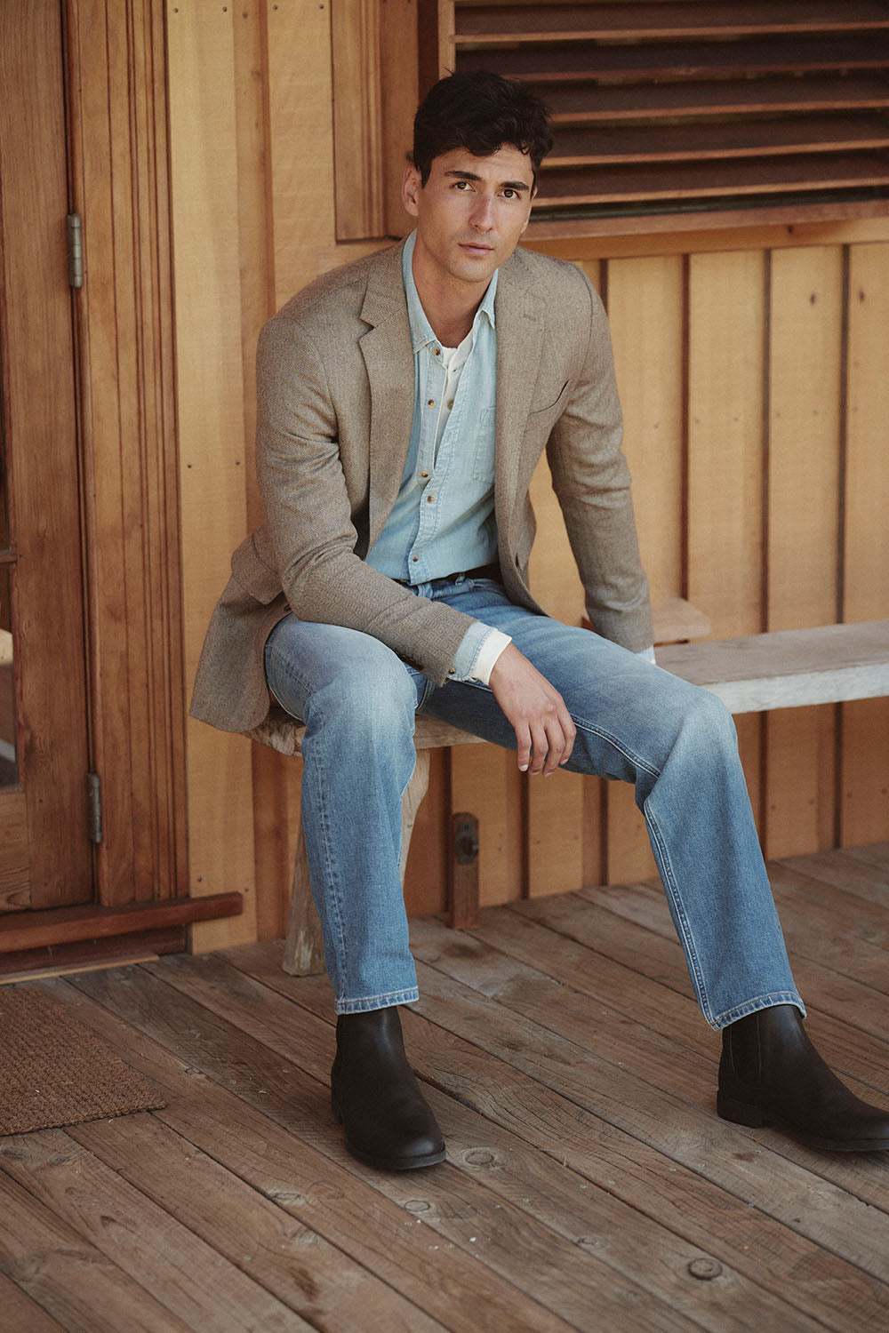 Man in a tan blazer, denim shirt, and jeans sits on a wooden bench outdoors in NZ.