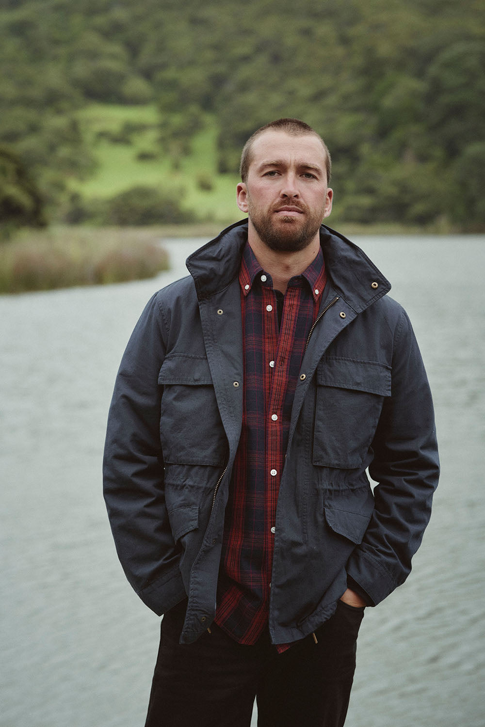Man in a navy jacket and plaid shirt stands by an NZ lake with trees in the background.