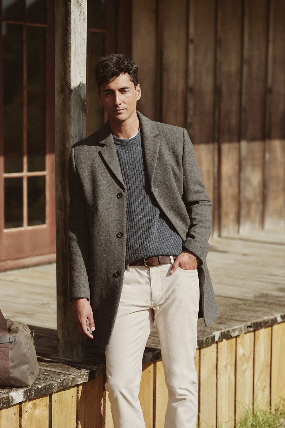 Man in a gray coat and beige pants leans against a wooden NZ porch railing outside.