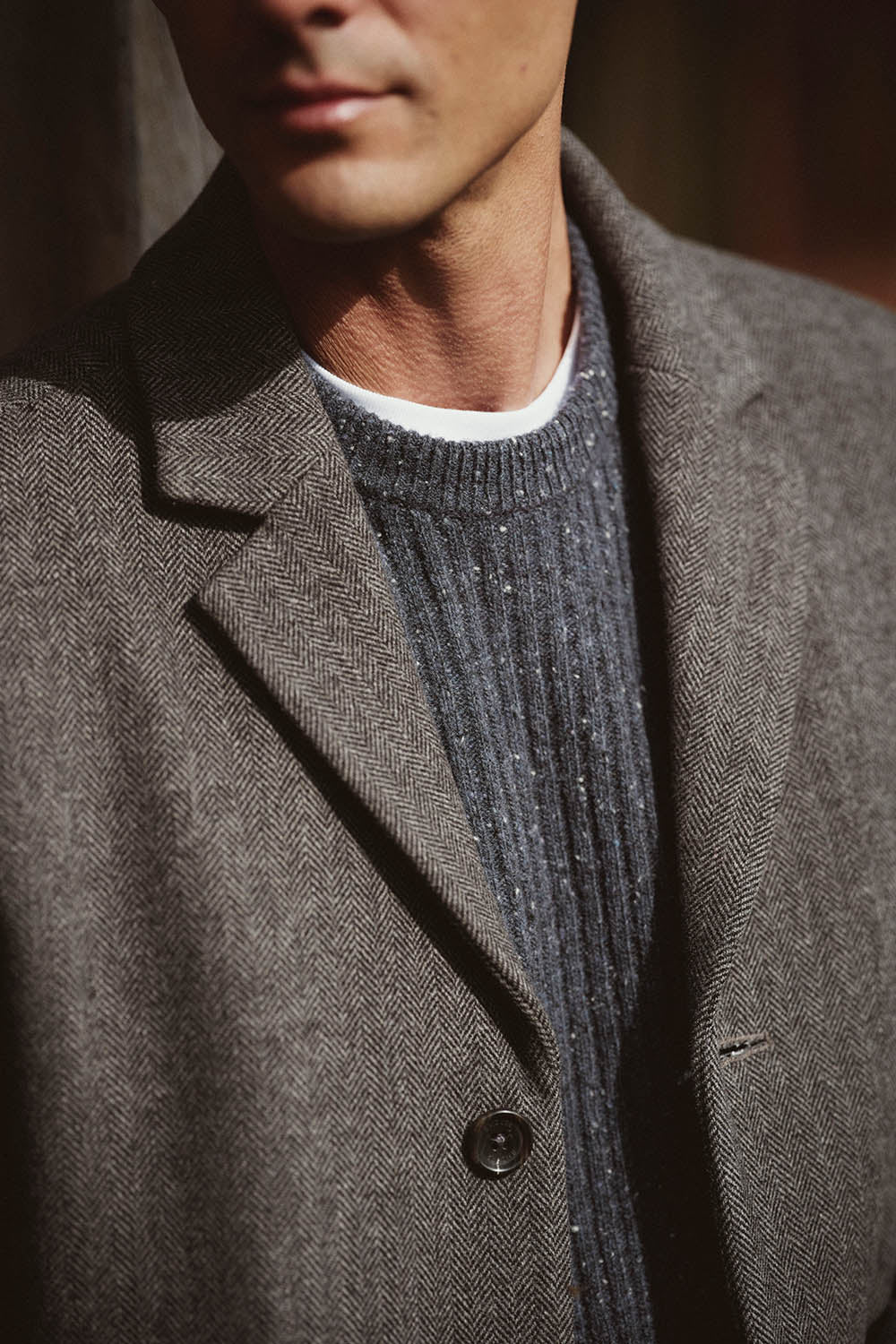Man wearing a grey herringbone coat, NZ style, over a dark ribbed sweater and white shirt.
