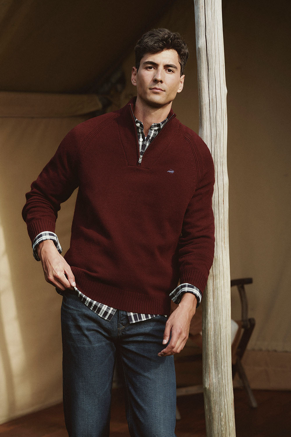 Man in a maroon sweater and jeans stands by a wooden post inside a canvas tent in NZ.