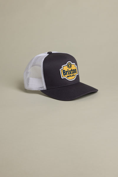 The Brixton International Trucker by Brixton is an eco-friendly black and white mesh trucker hat with a yellow and black patch on the front, shown against a neutral beige background.