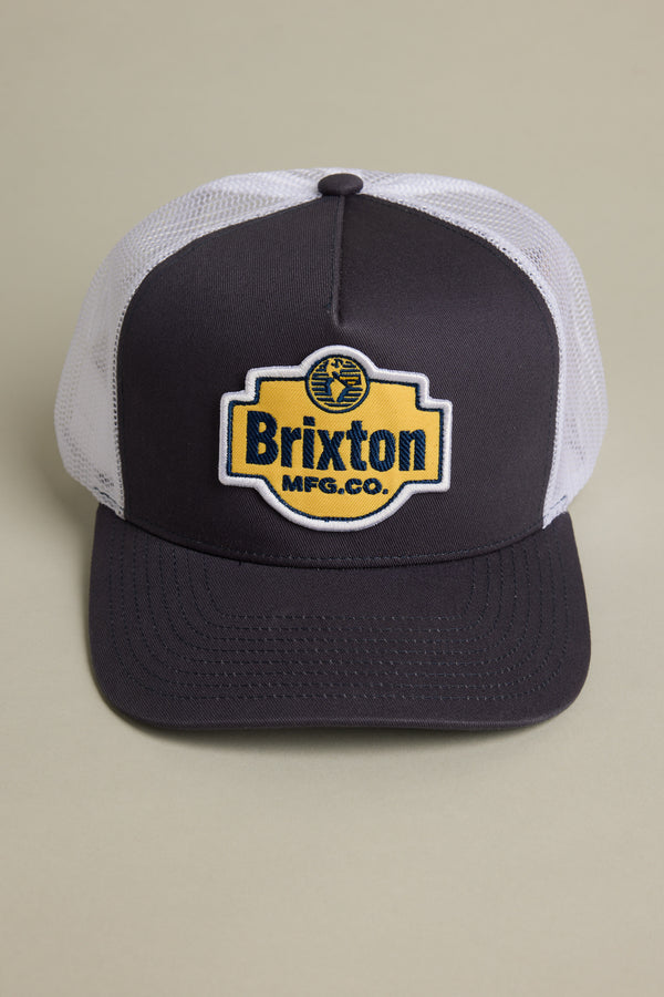 The Brixton International Trucker by Brixton is a black and white mesh hat made from recycled fishing nets, featuring a yellow and black patch on the front—a stylish, eco-friendly statement. Displayed on a light beige background.