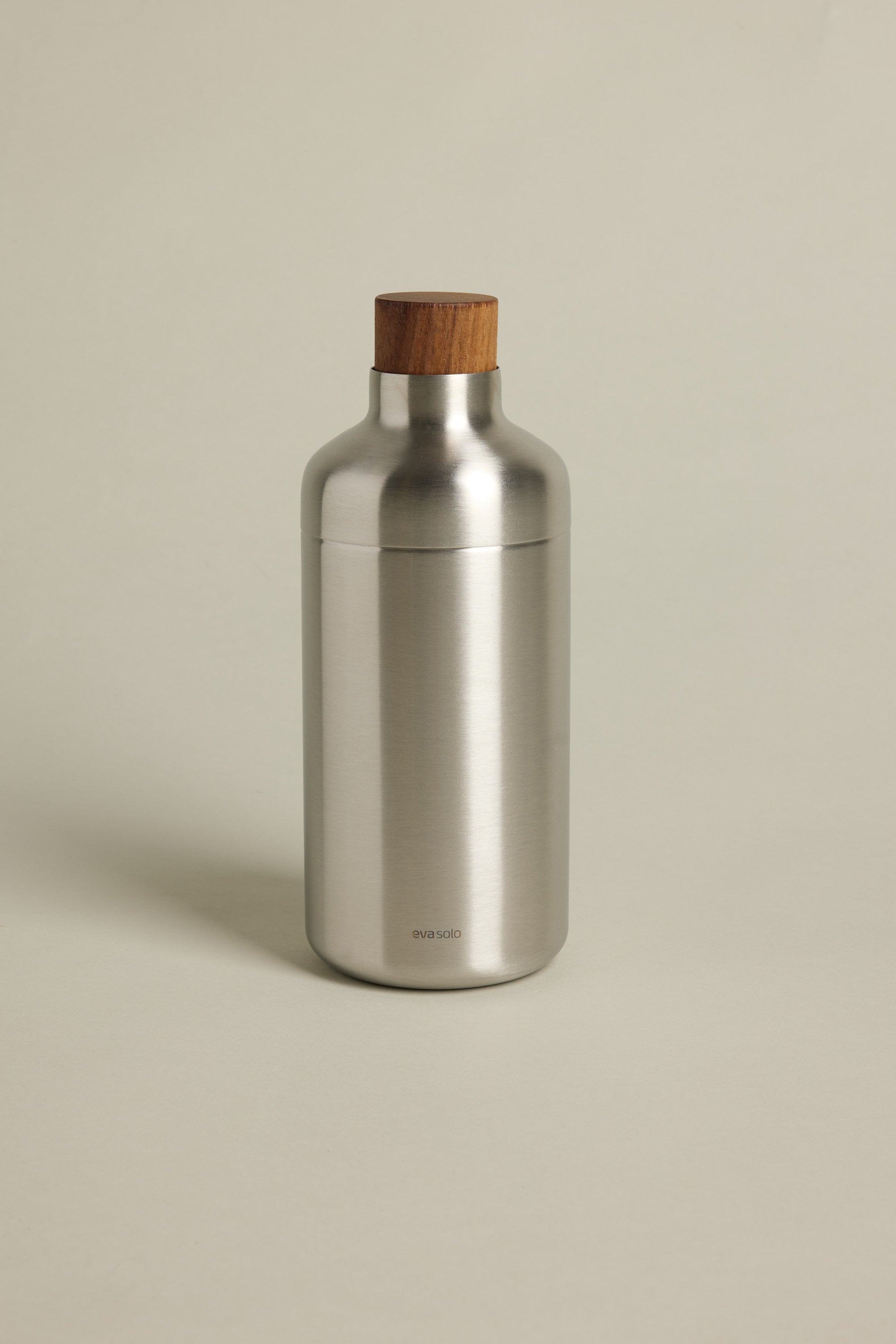 The Other Liquid Lounge Cocktail Shaker, featuring a stainless steel body and wooden cap, stands upright on a light beige background. Its minimalistic, modern design with engraved logo makes it a must-have for stylish home bar enthusiasts.