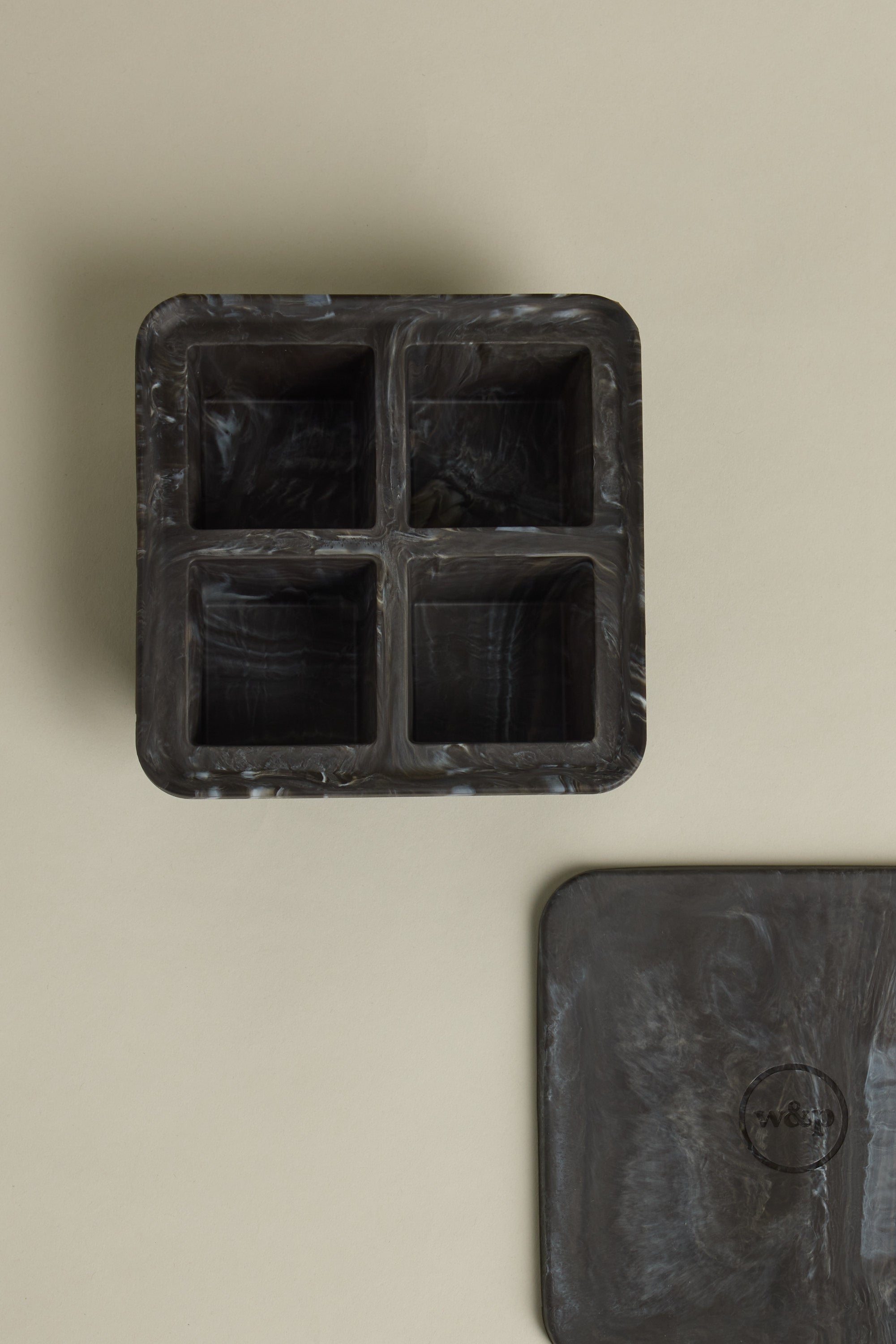 The Other Ice Tray Sphere Marble Black, featuring a black and gray marble look with four square compartments, is shown on a beige surface. Its matching lid with a circular logo appears in the lower right corner. Perfect for any home bar.