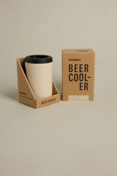 A cream Huski Beer Cooler with a black lid is shown in a cardboard holder beside its matching brown box labeled "Huski Beer Cooler." Both are on a beige background, highlighting triple-insulated stainless steel for high-performance cooling.