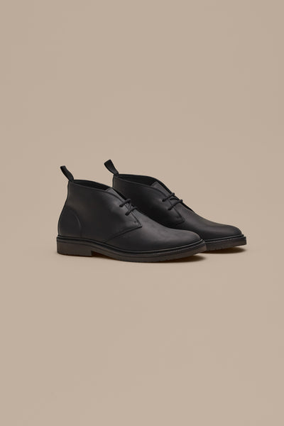 The Barkers Howarth Desert Boot, a minimalist black leather chukka inspired by classic desert boots, sits side by side on a light beige background, featuring laces, pull tabs at the back, and a slight heel.