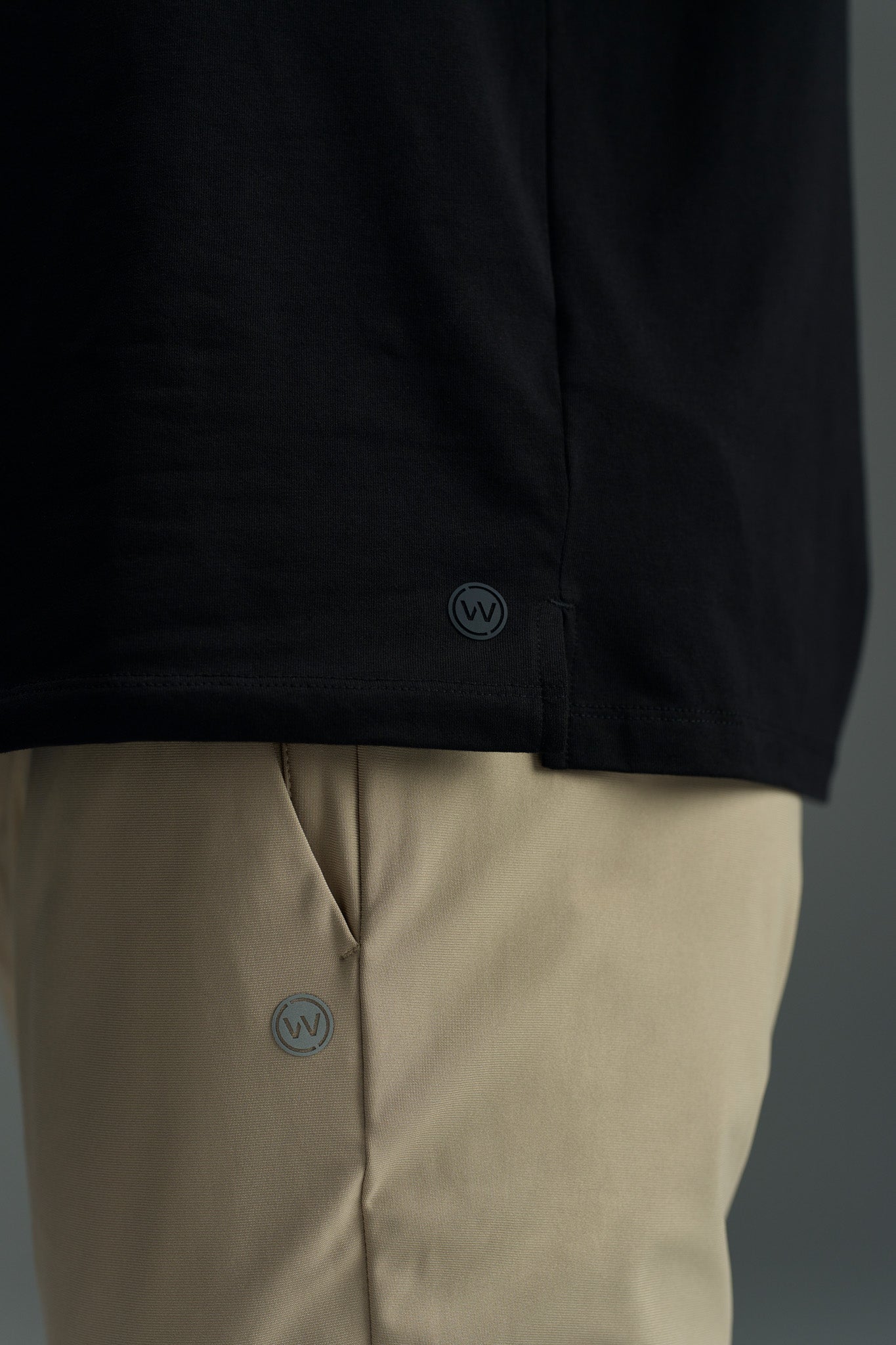 A close-up of someone wearing a black shirt and beige WearEver Long Game Pants, both marked with WearEver’s small circular WV logo near the hem and pocket.