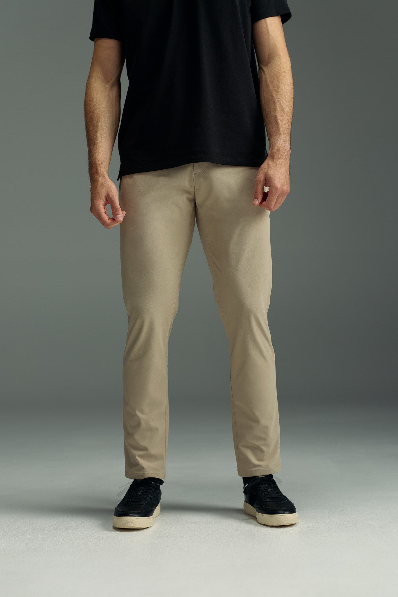 A person stands against a plain background, wearing a black t-shirt, black sneakers, and beige WearEver Long Game Pants by WearEver. The image is cropped at the shoulders, so the person's face is not visible.