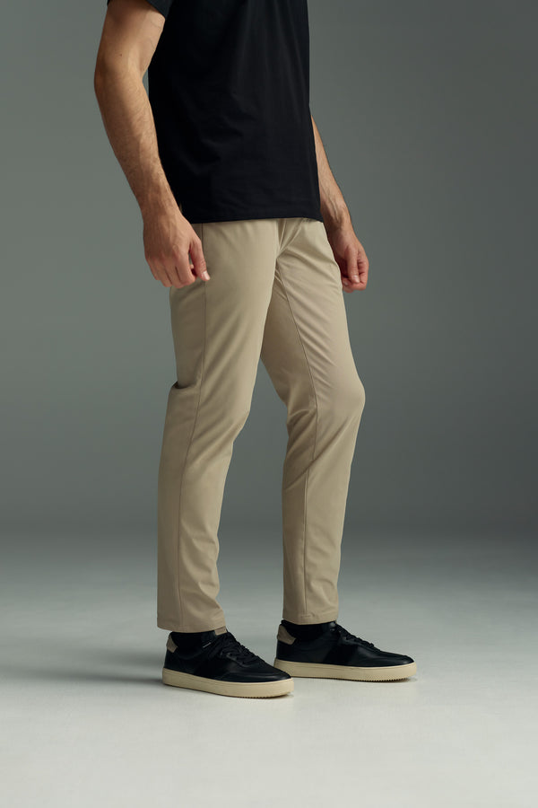 A person stands on a light gray floor wearing the WearEver Long Game Pant in beige, a black t-shirt, and black sneakers with light-colored soles. The upper part of the face is not visible.