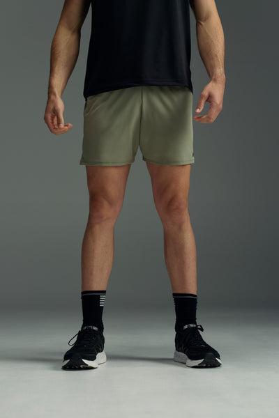 A person stands with arms relaxed at their sides, wearing a black T-shirt and WearEver Cadence Short lightweight running shorts with antibacterial treatment, paired with black socks and athletic shoes, against a plain gray background. Face not visible.